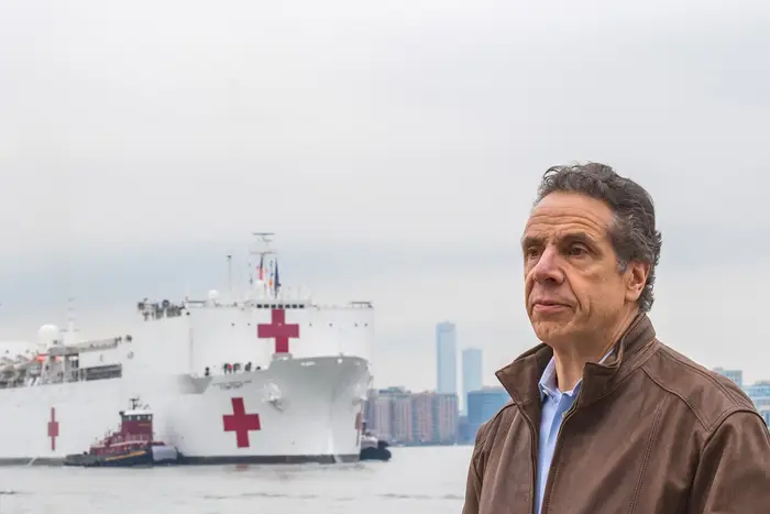 Governor Cuomo stands on a pier, with the USNS Comfort hospital ship, which is white with red crosses on it, in the river behind him